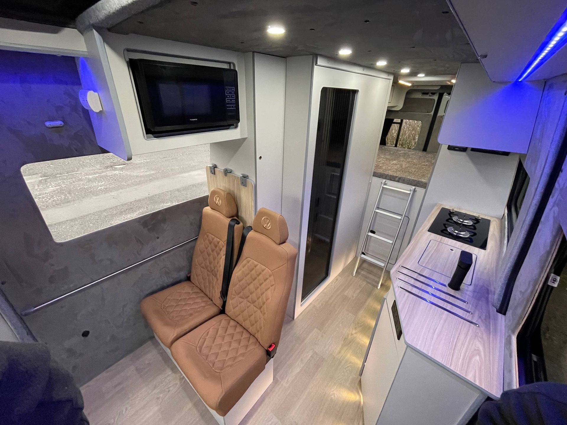 Compact modern RV interior with tan seats, wall-mounted TV, and a small kitchen walkway.