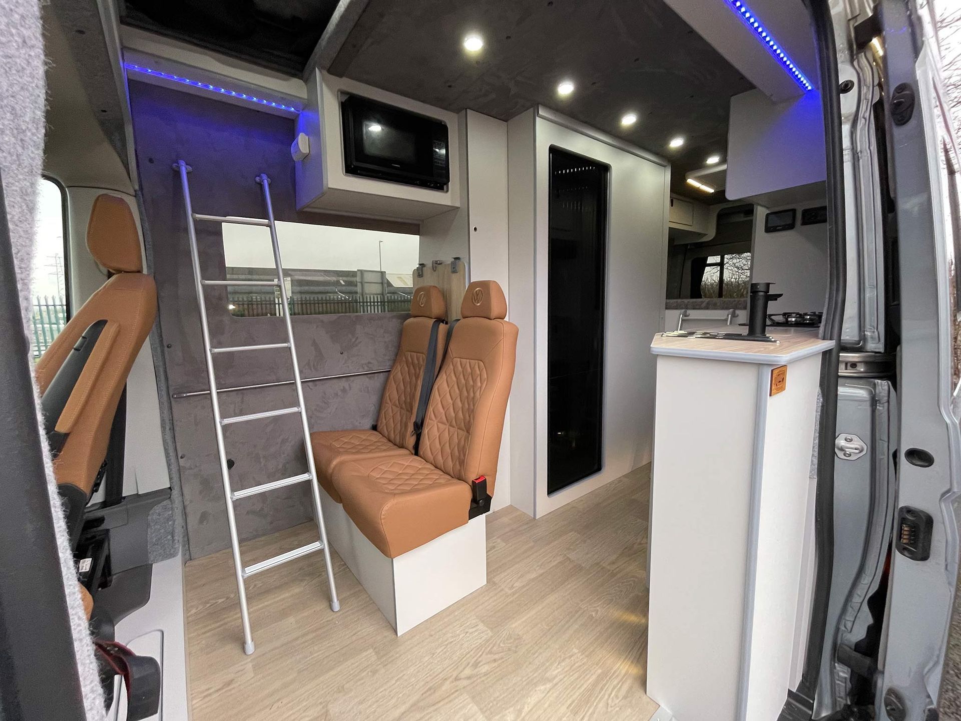 Modern camper van interior with tan bench seats, ladder, skylights, and a white counter.