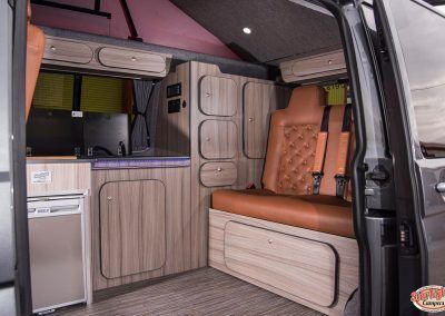 Compact camper van interior with wood cabinetry, bench seat, and bed storage under a pink roof
