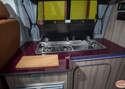 Compact camper kitchen with sink, stove, cutting board, and wood cabinetry