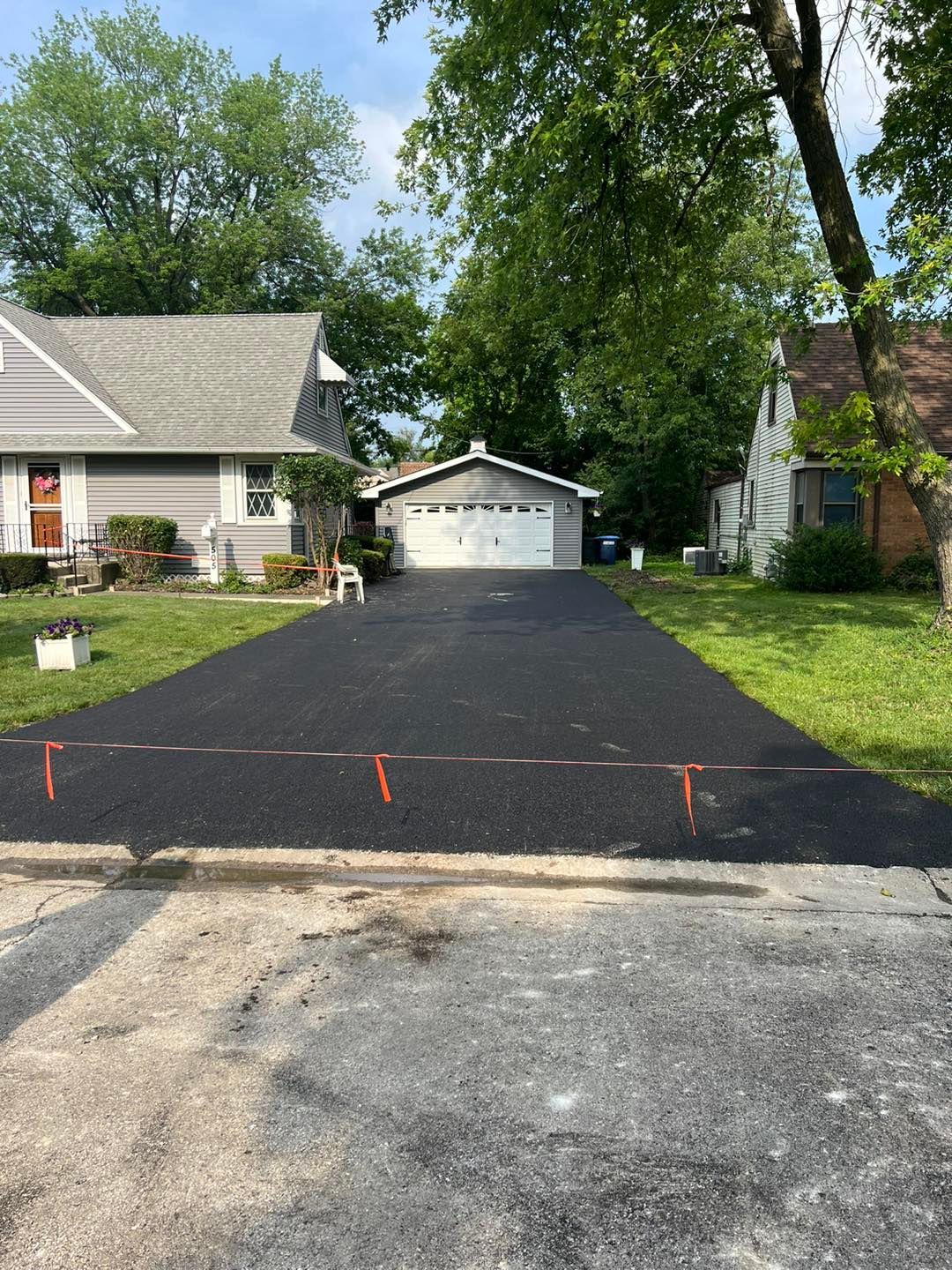Freshly paved black driveway leading to a white garage between two houses on a leafy street.