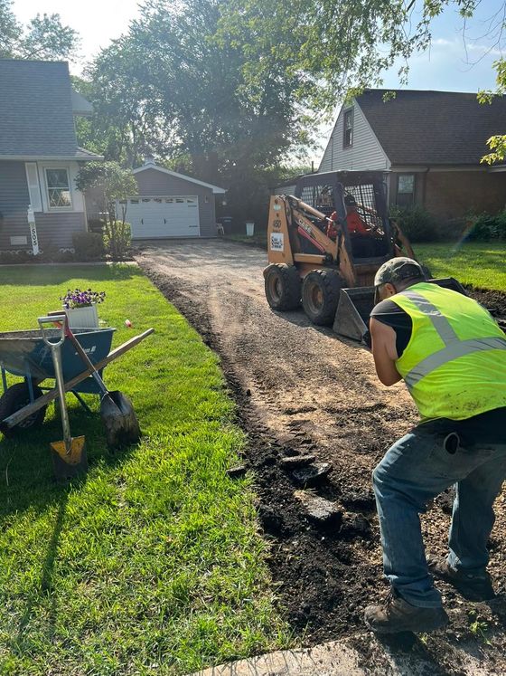 Worker using a skid steer to spread gravel along a driveway beside a grassy yard and house