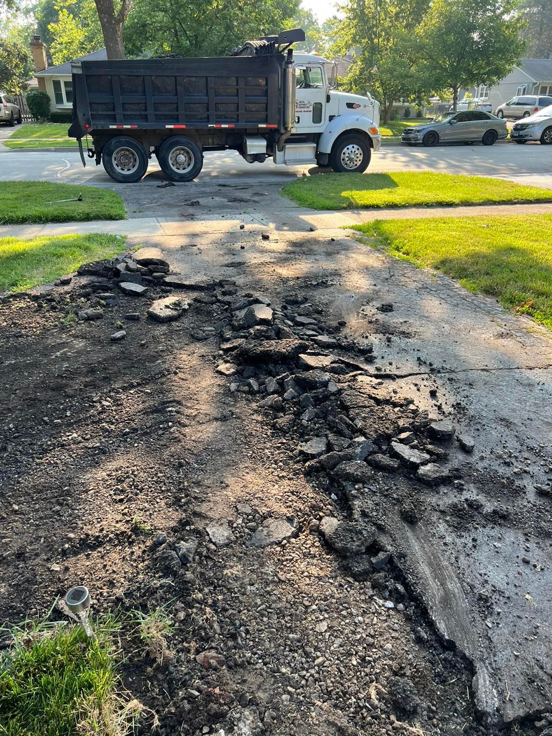 Cracked, patched asphalt road with a dump truck in the background on a sunny residential street.