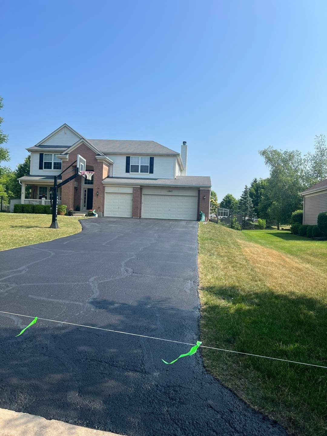 Suburban two-story house with brick facade, double garage, and a long blacktop driveway under a clear blue sky