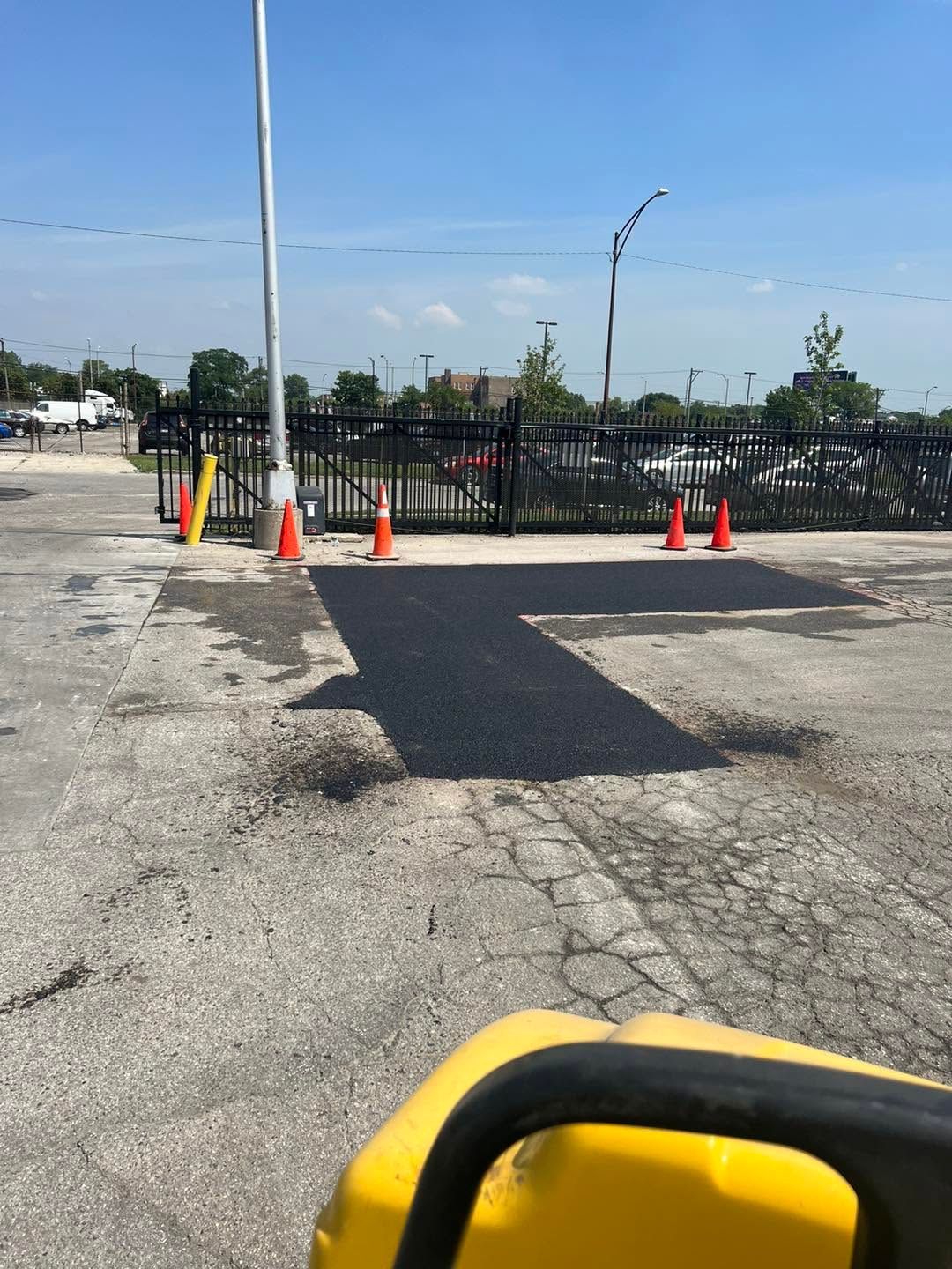 Parking lot with fresh black asphalt patch, orange cones, and a metal fence under a clear blue sky
