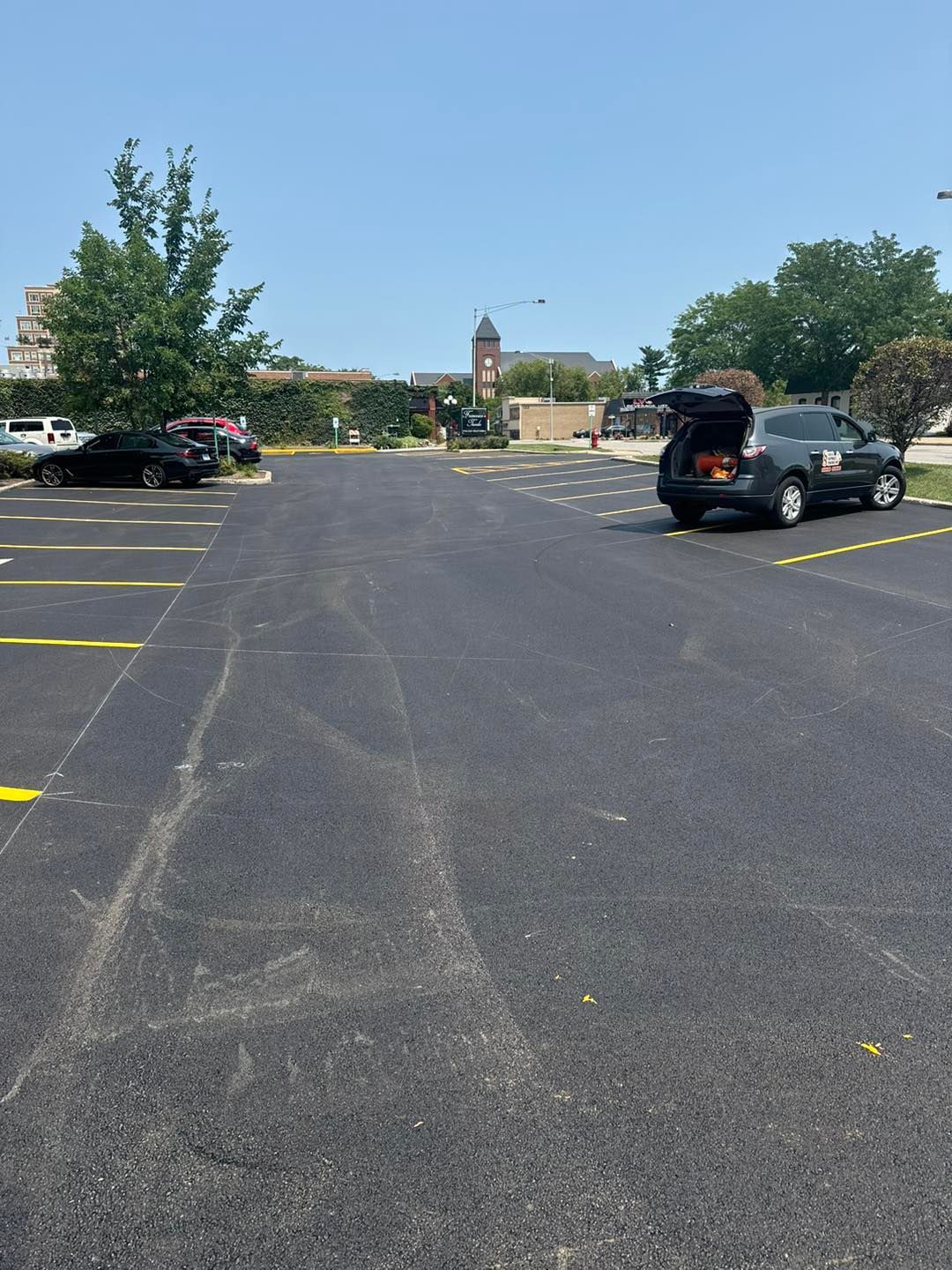 Mostly empty asphalt parking lot with a few cars, trees, and a distant building under a blue sky