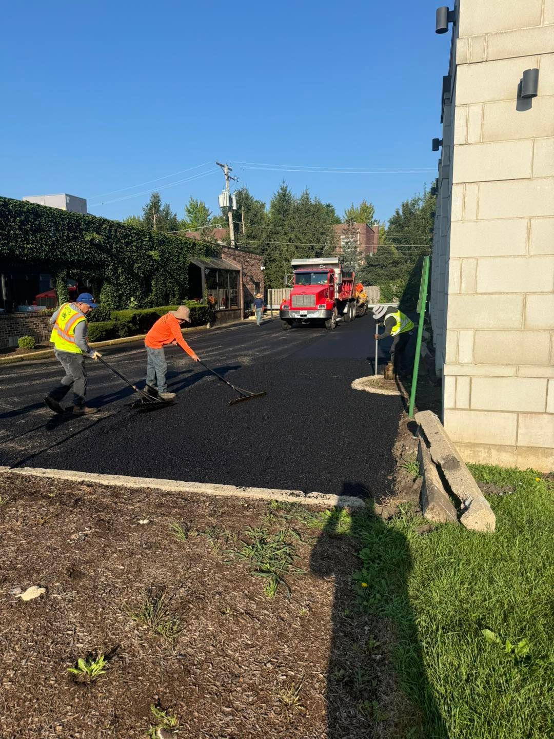 Workers paving a driveway with fresh black asphalt beside a building, with a red truck in the background.