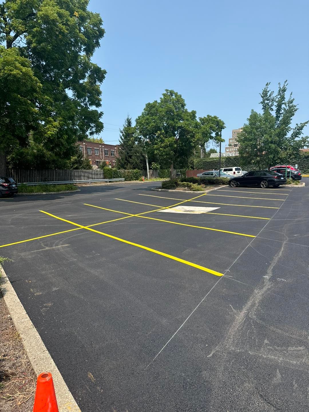 Empty asphalt parking lot with yellow lines, trees, and parked cars under a clear blue sky