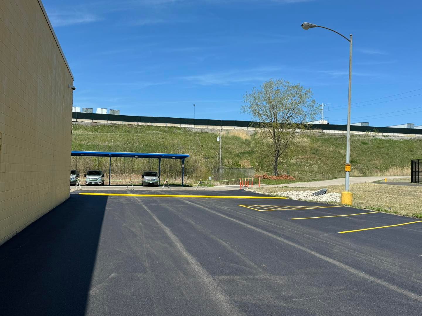 Empty parking lot beside a beige building, with a bus stop shelter and grassy embankment under blue sky