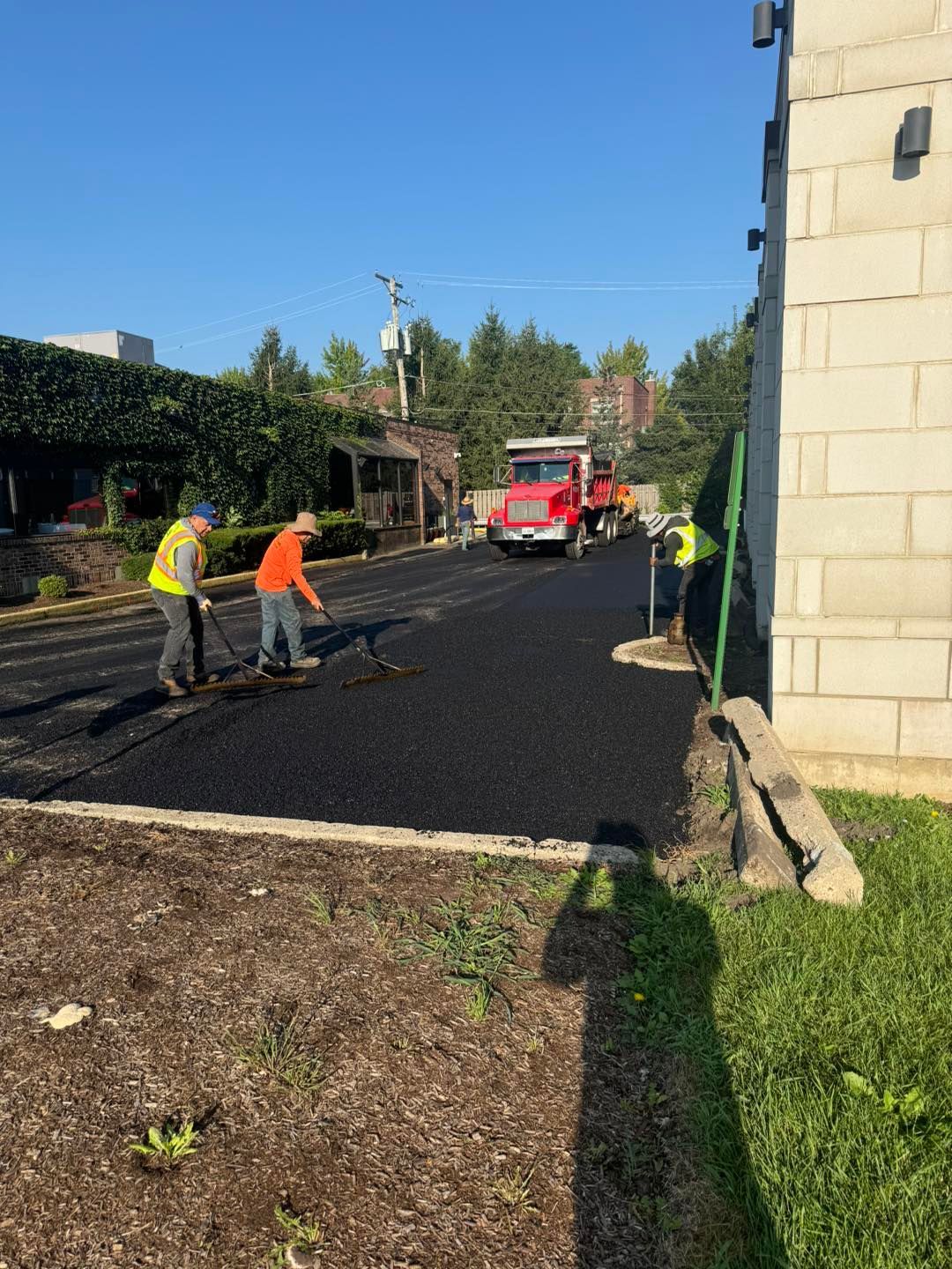 Road crew paving a street, with workers, a red truck, and fresh black asphalt.