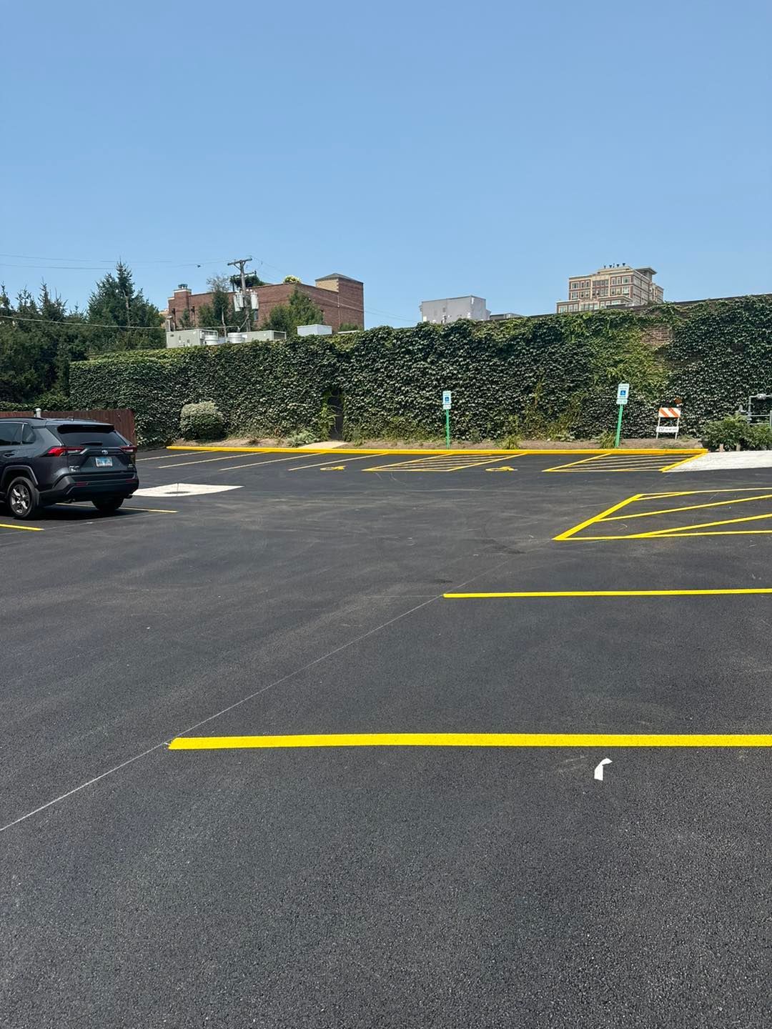 Empty asphalt parking lot with yellow painted lines, cars at left, and a vine-covered wall in back