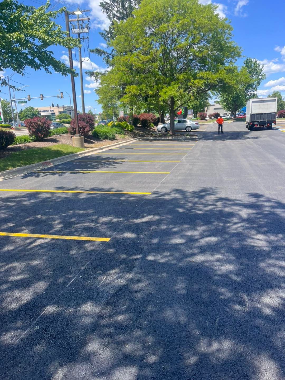 Sunny parking lot with yellow lines, trees, a few people, and a white truck on the road.