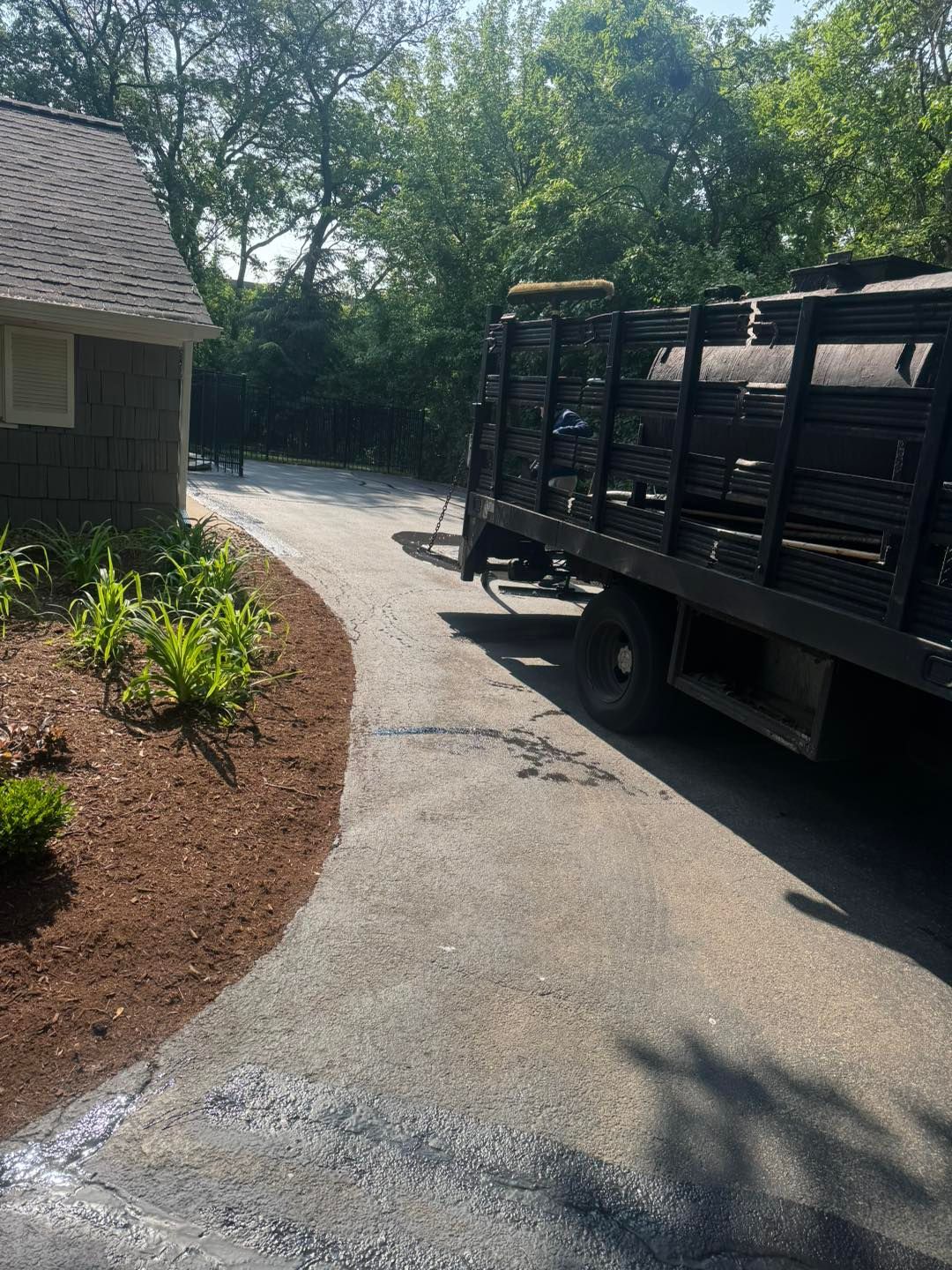 Driveway beside a house with a parked black utility trailer and sunny trees in the background
