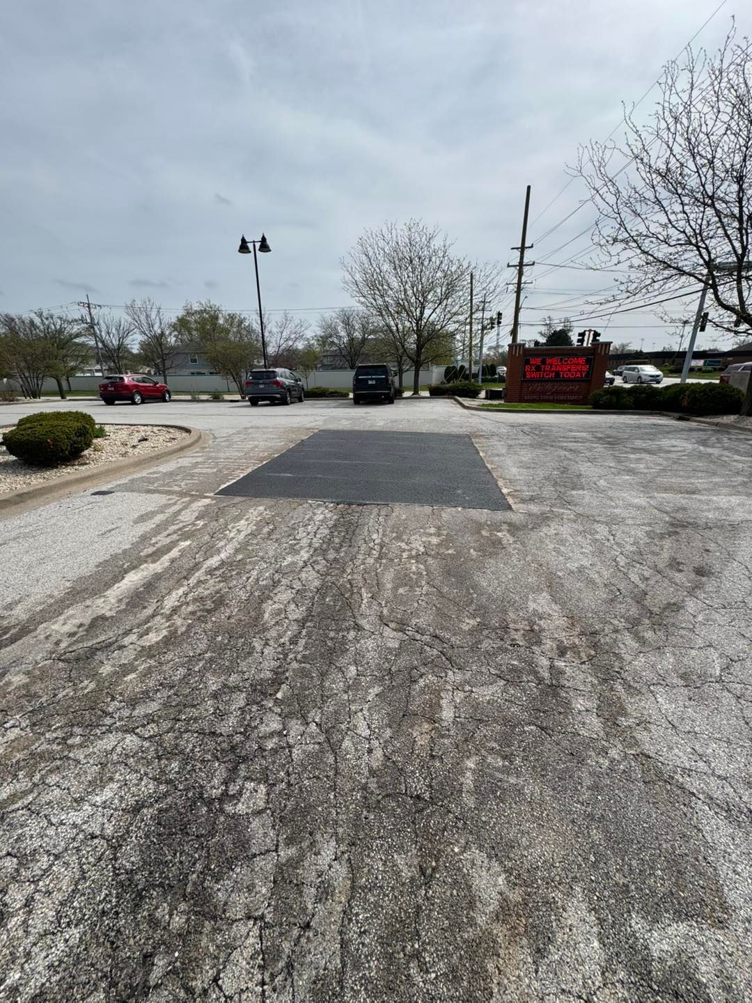 Cracked asphalt lot with parked cars, a red sign, leafless trees, and cloudy sky