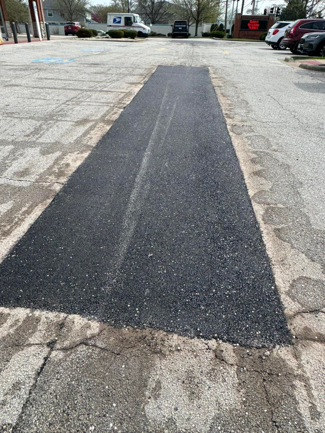 Fresh black asphalt patch in a parking lot, surrounded by cracked pavement and parked cars.