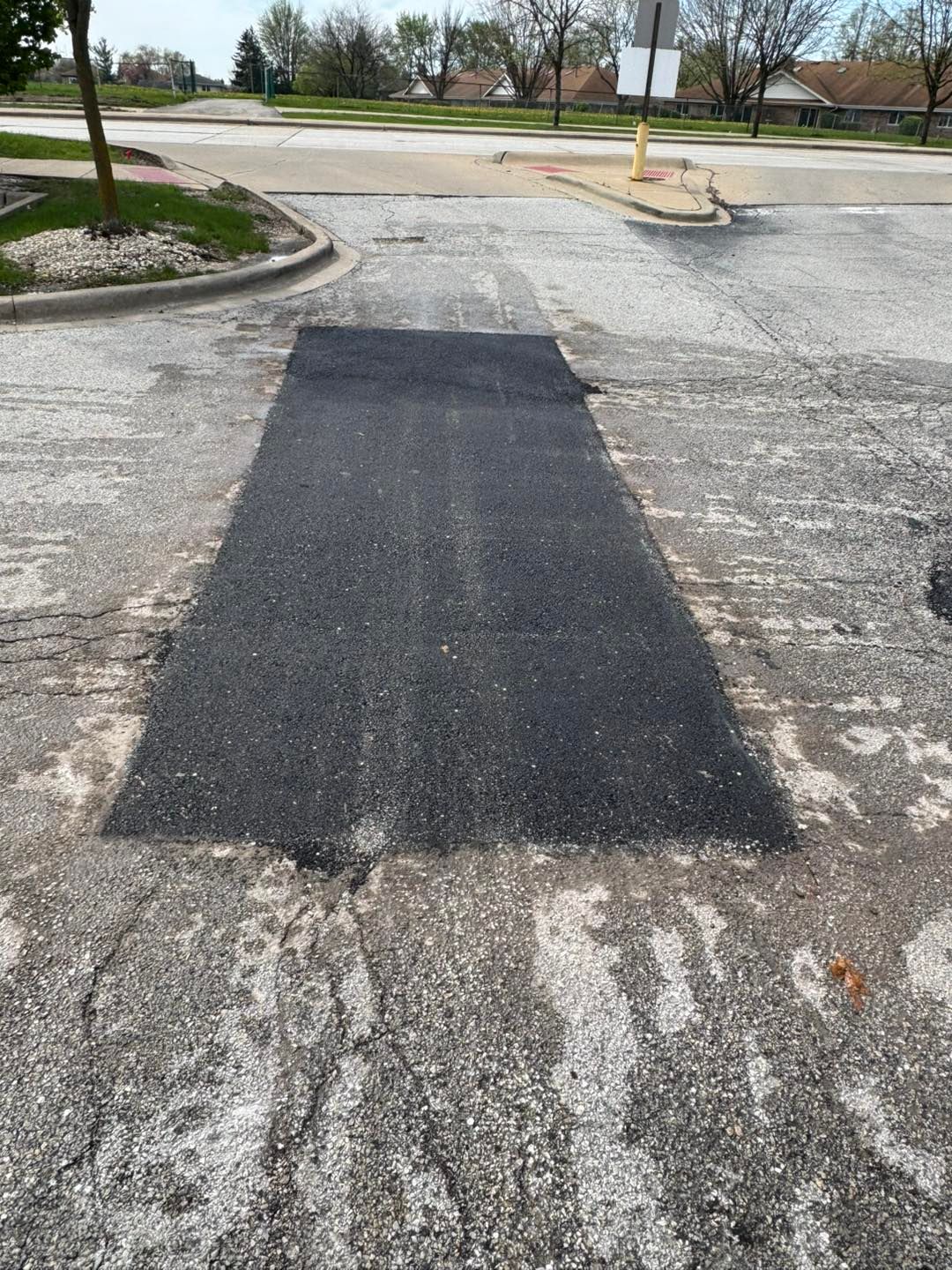 Fresh black asphalt patch on a cracked parking lot near a curb and grassy area.