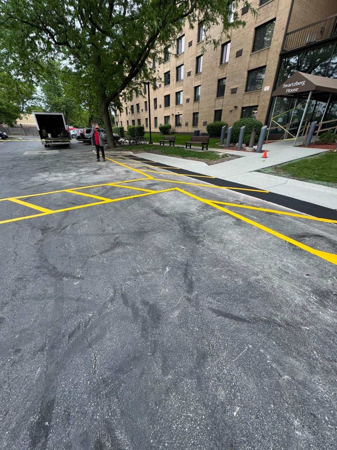 Apartment building entrance with yellow crosswalk markings on a tree-lined street, pedestrians in the distance