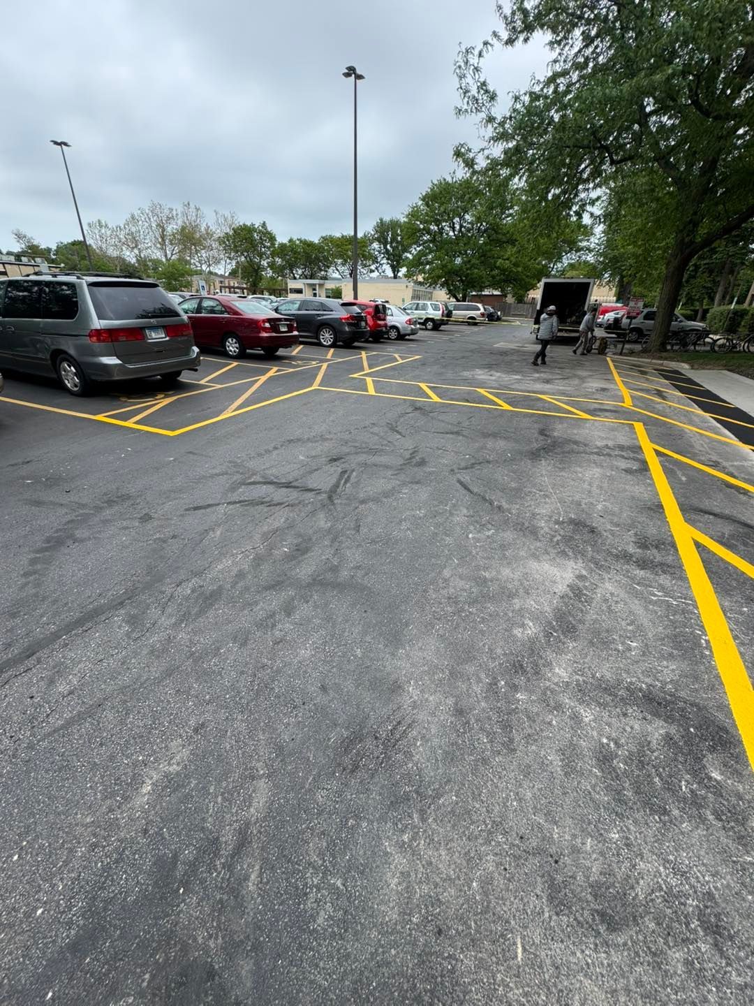 Parking lot with yellow striped markings, parked cars, and trees under an overcast sky