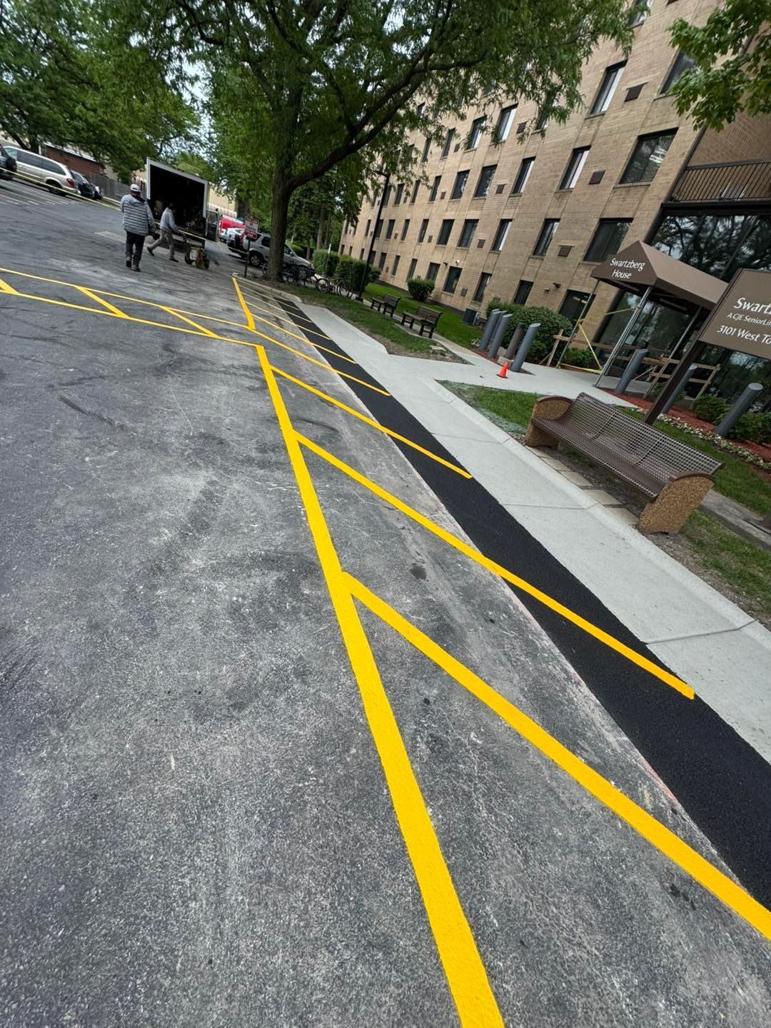 Slanted street with yellow lane markings beside an apartment building and sidewalk tree.
