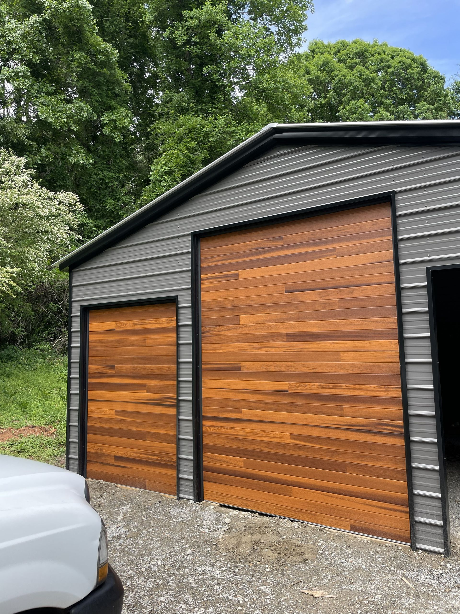Gray metal garage with two brown wood garage doors, gravel driveway, and green trees.