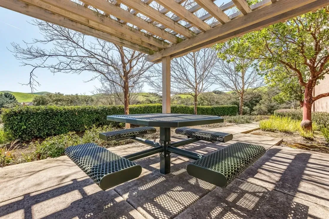 Picnic table under a pergola with a view of rolling hills and manicured hedges.