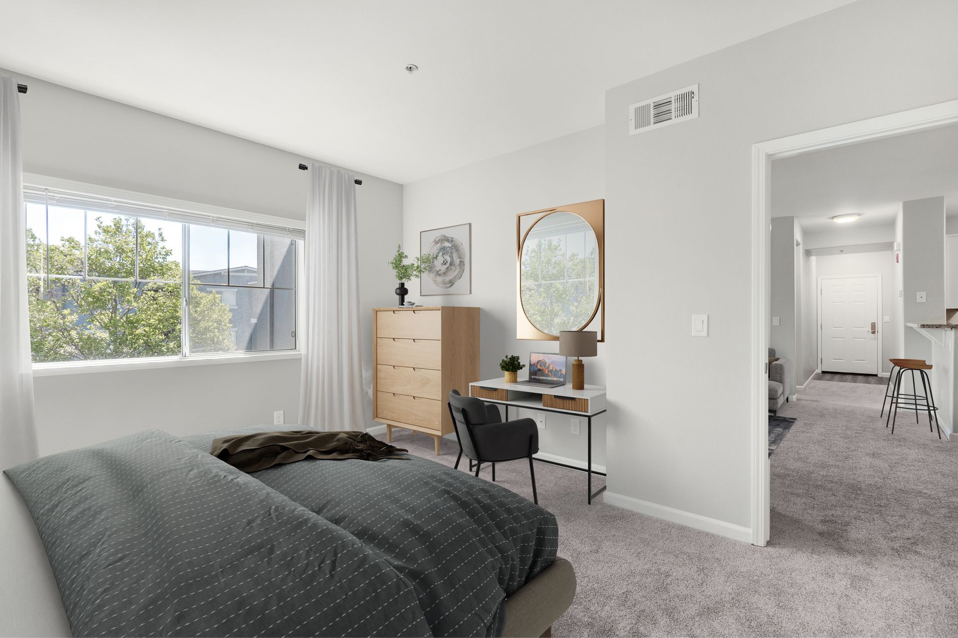 Primary bedroom with a large window, desk workspace, dresser, and plush carpeting overlooking the apartment entryway.
