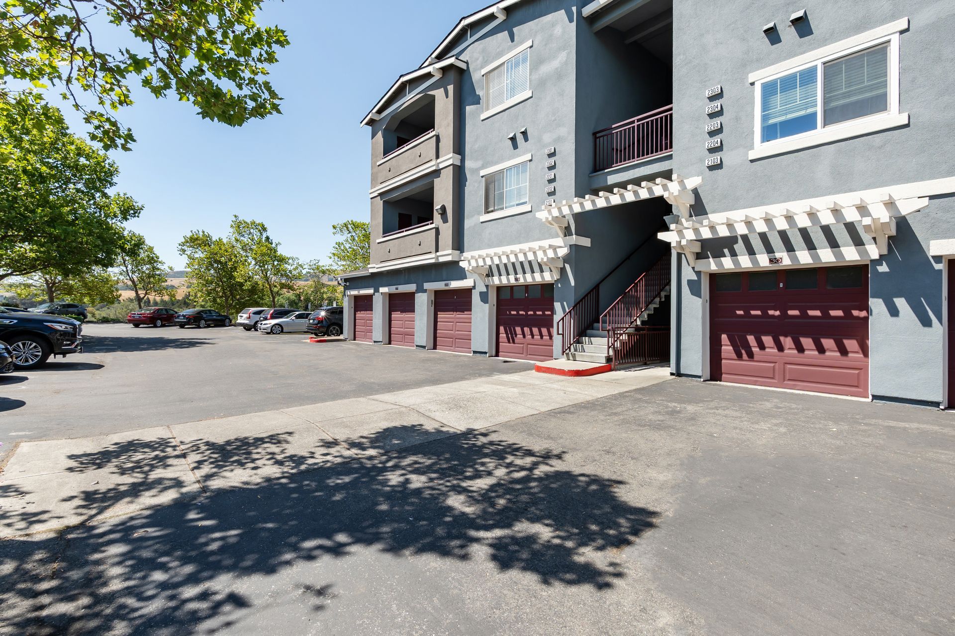 Exterior of apartment community with private garages, covered balconies, on-site parking, and mature trees.