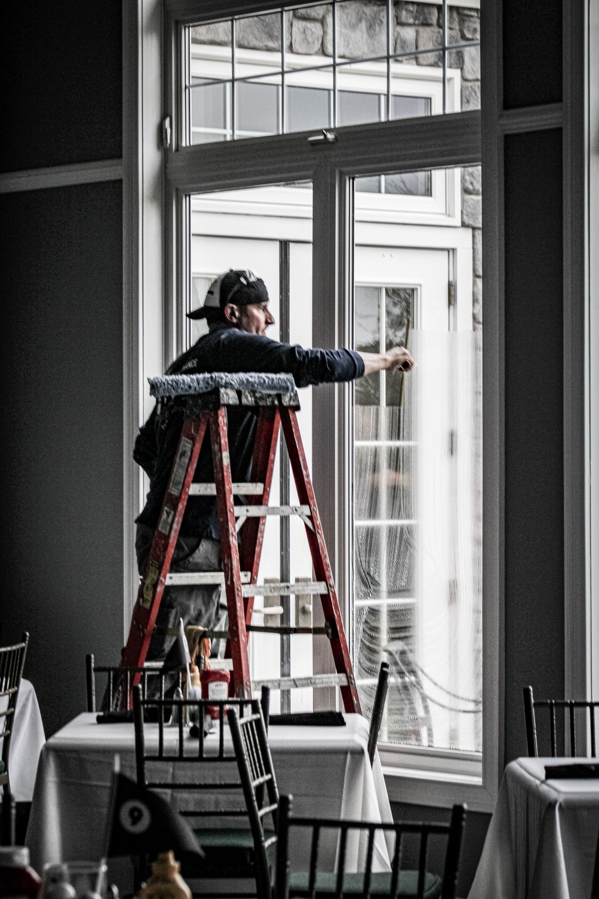 A man is standing on a ladder cleaning a window in a restaurant.