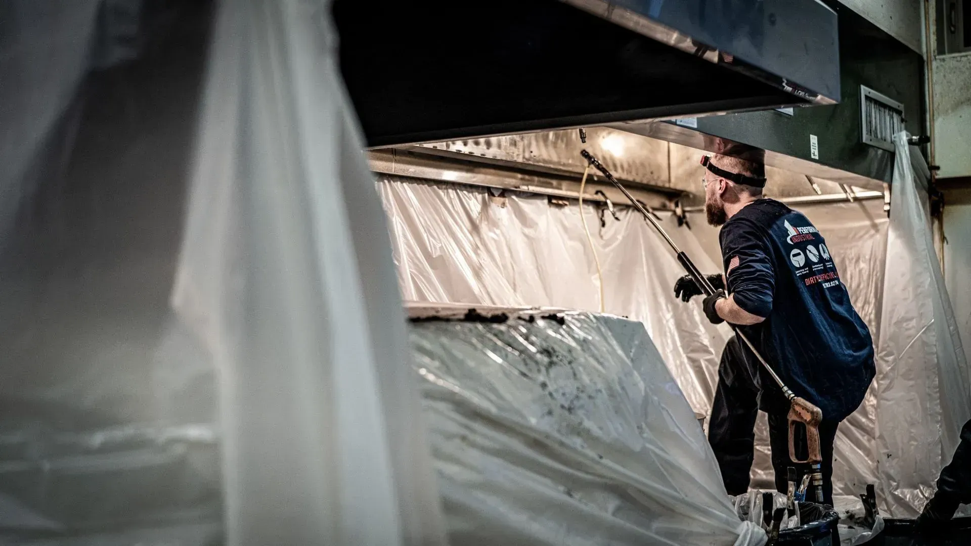 Kitchen exhaust cleaning technician spraying commercial hood system safely wrapped in plastic.