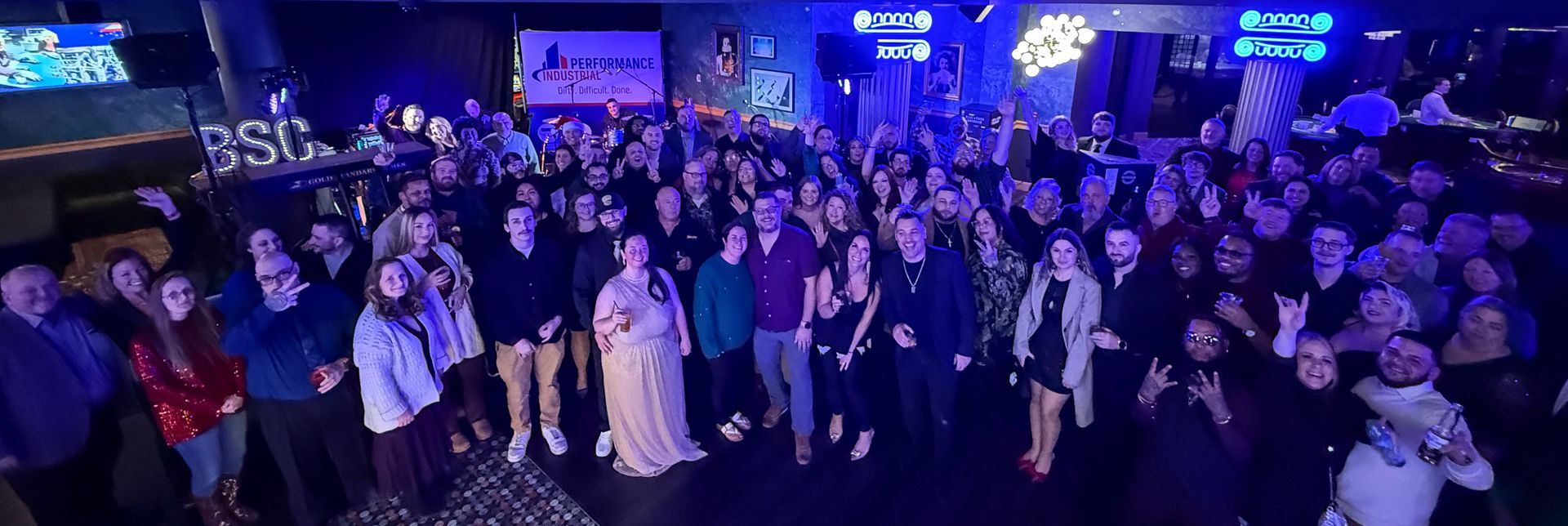 A large group of Performance Industrial employees posing together and celebrating at the company's Christmas party in December 2025, held in the rented Rec Room at Turning Stone Casino, with blue stage lighting, a banner, and people raising drinks and making hand signs in a festive atmosphere that included live music from Big Sky, raffles, and hotel accommodations provided for the team. 