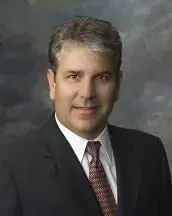 Man in suit and tie, smiling at the camera, against a dark, blurred background.