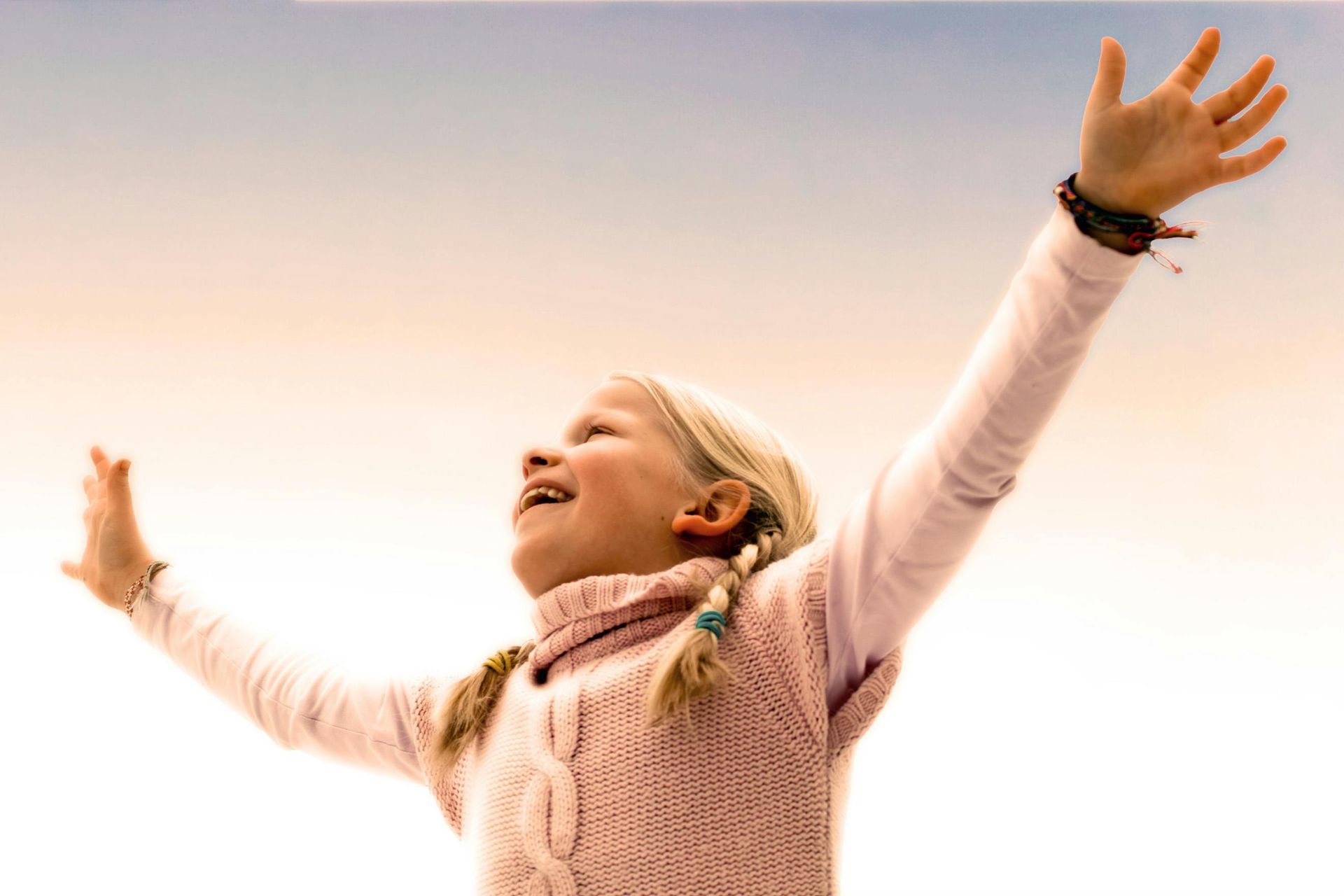 Girl with blonde braids, arms raised, looking up with joyful expression against a bright sky.