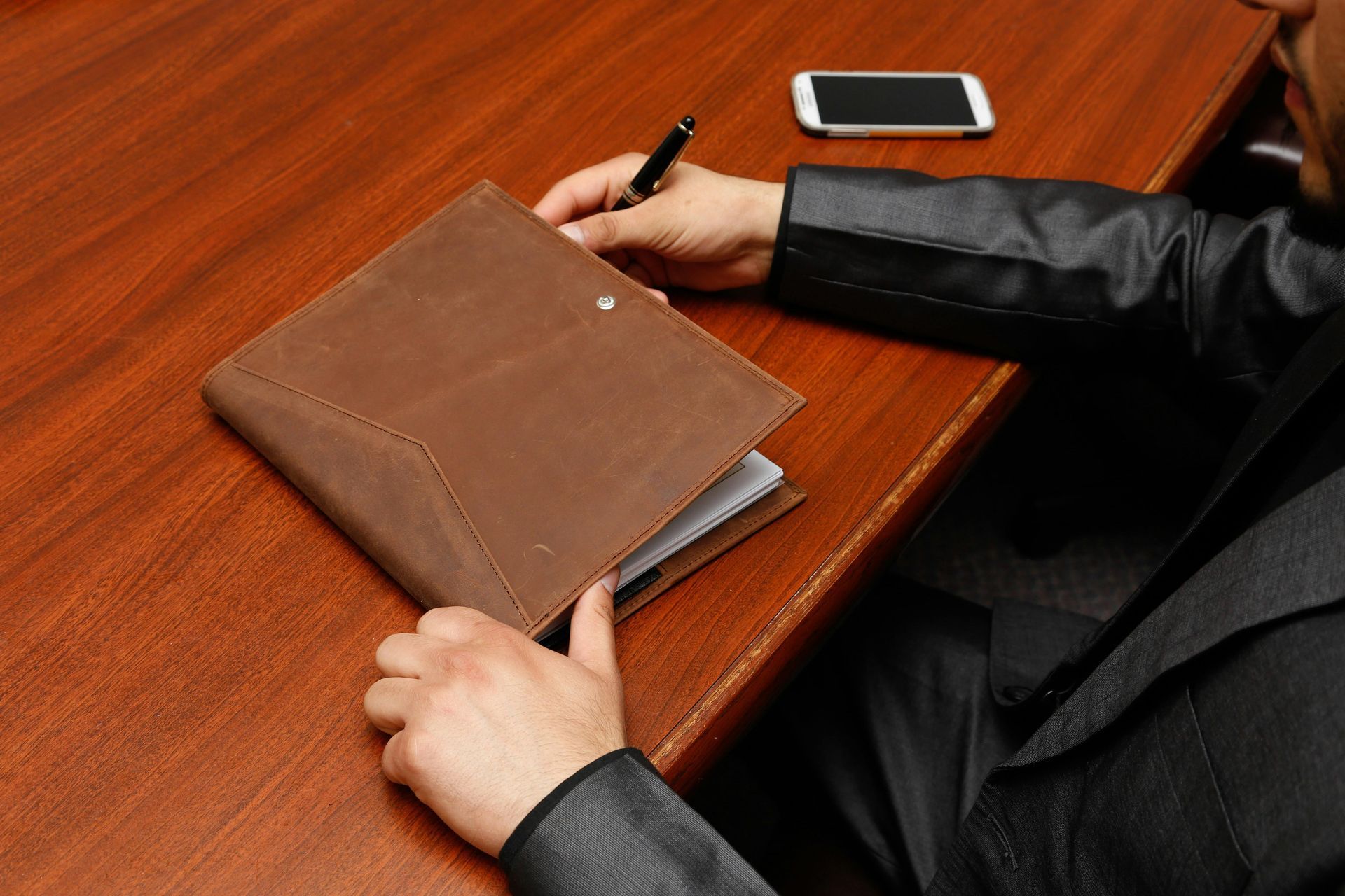 Person in a black suit opening a brown leather notebook on a wooden desk next to a smartphone.