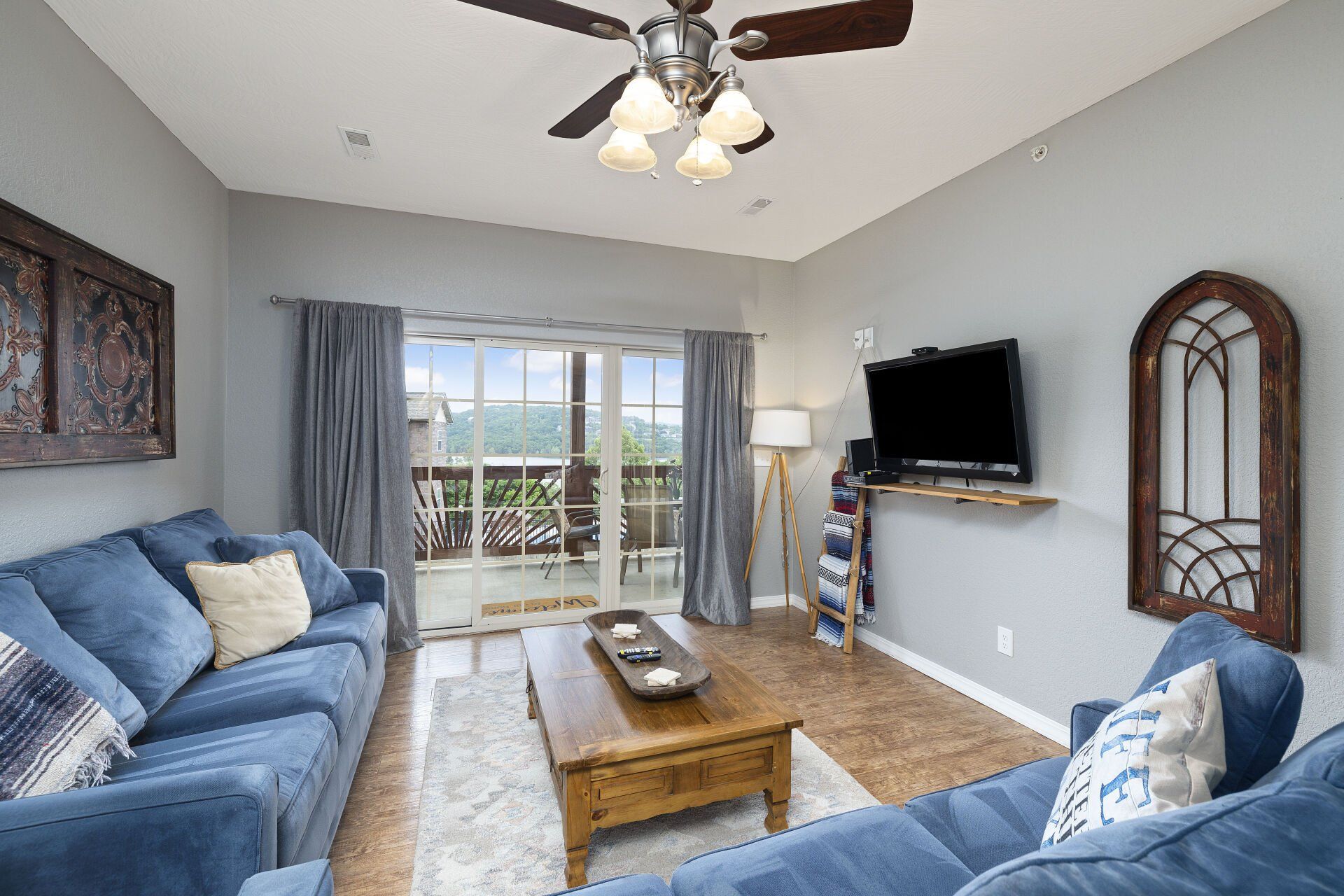 Bright living room with blue sofas, coffee table, TV, and balcony doors overlooking greenery