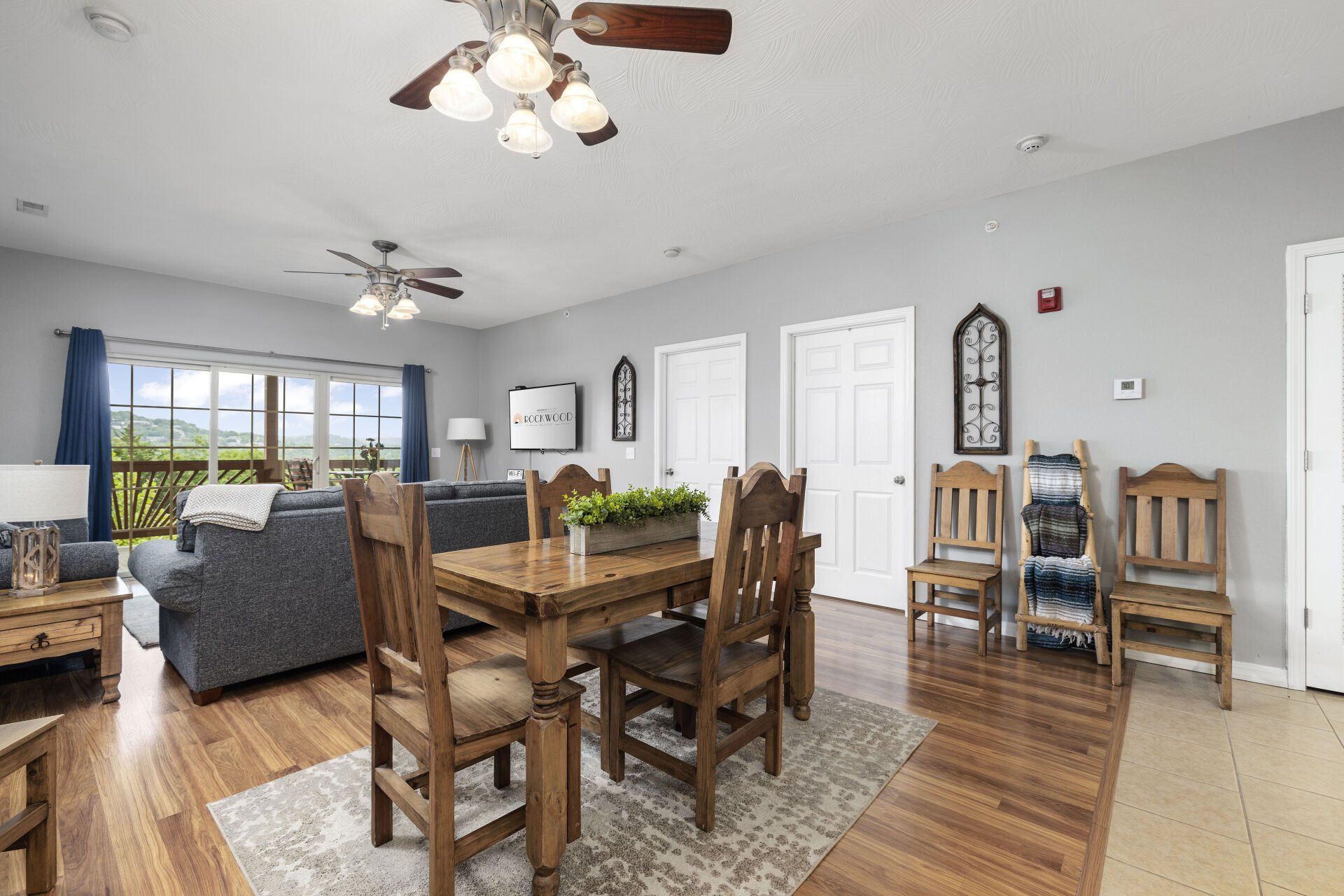 Open dining and living room with wood table, gray sofa, ceiling fans, and large window view