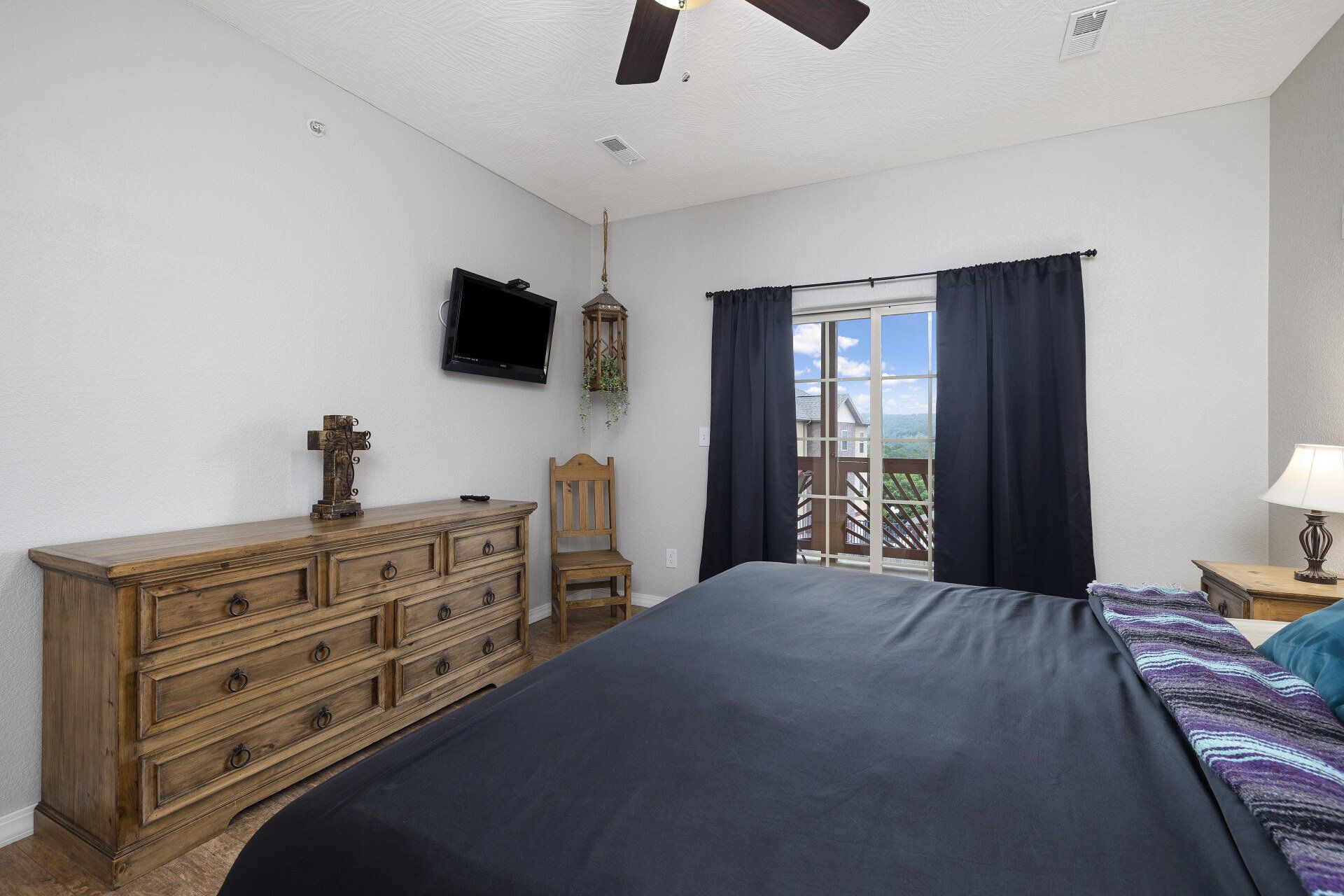Bedroom with dark bedspread, wooden dresser, wall-mounted TV, and sliding doors with curtains.