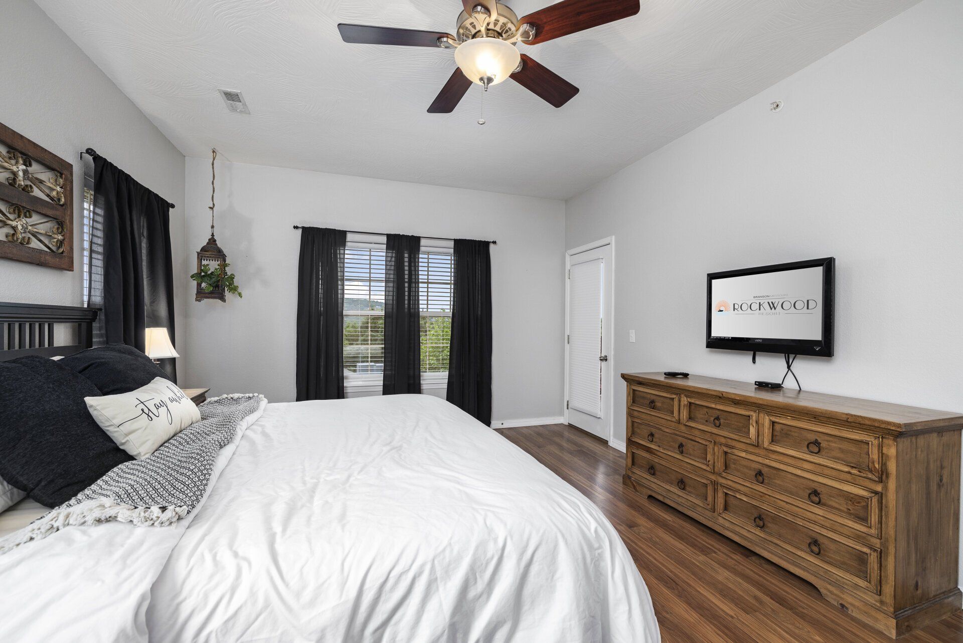 Bright bedroom with a bed, dark curtains, ceiling fan, and wooden dresser with a TV