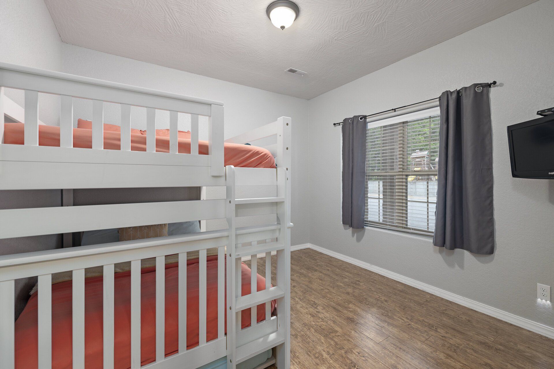 Children’s bedroom with white bunk bed, orange bedding, gray curtains, and wall-mounted TV