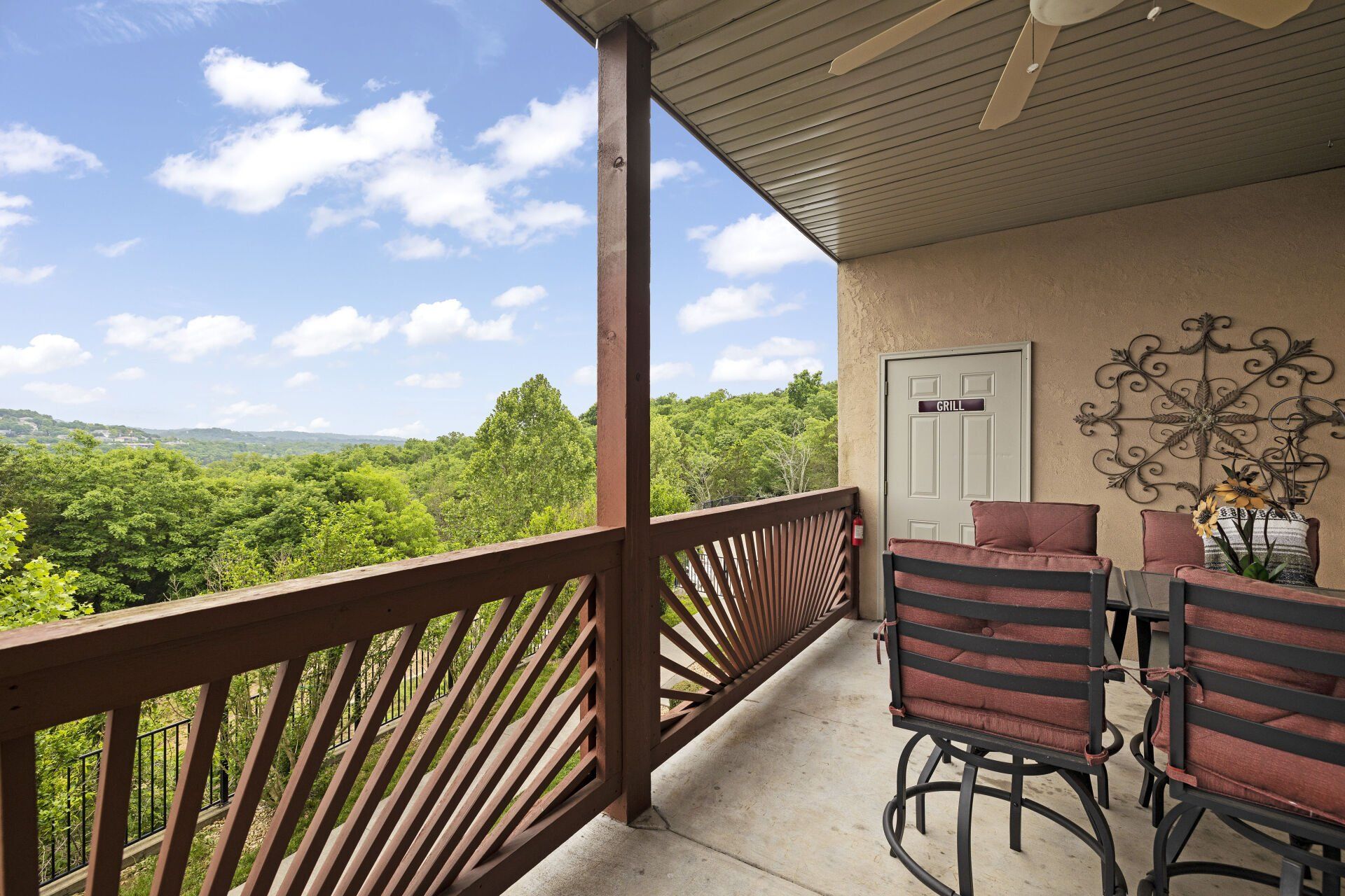 Covered balcony with patio table, striped chairs, wood railing, and a view of green trees and blue sky.