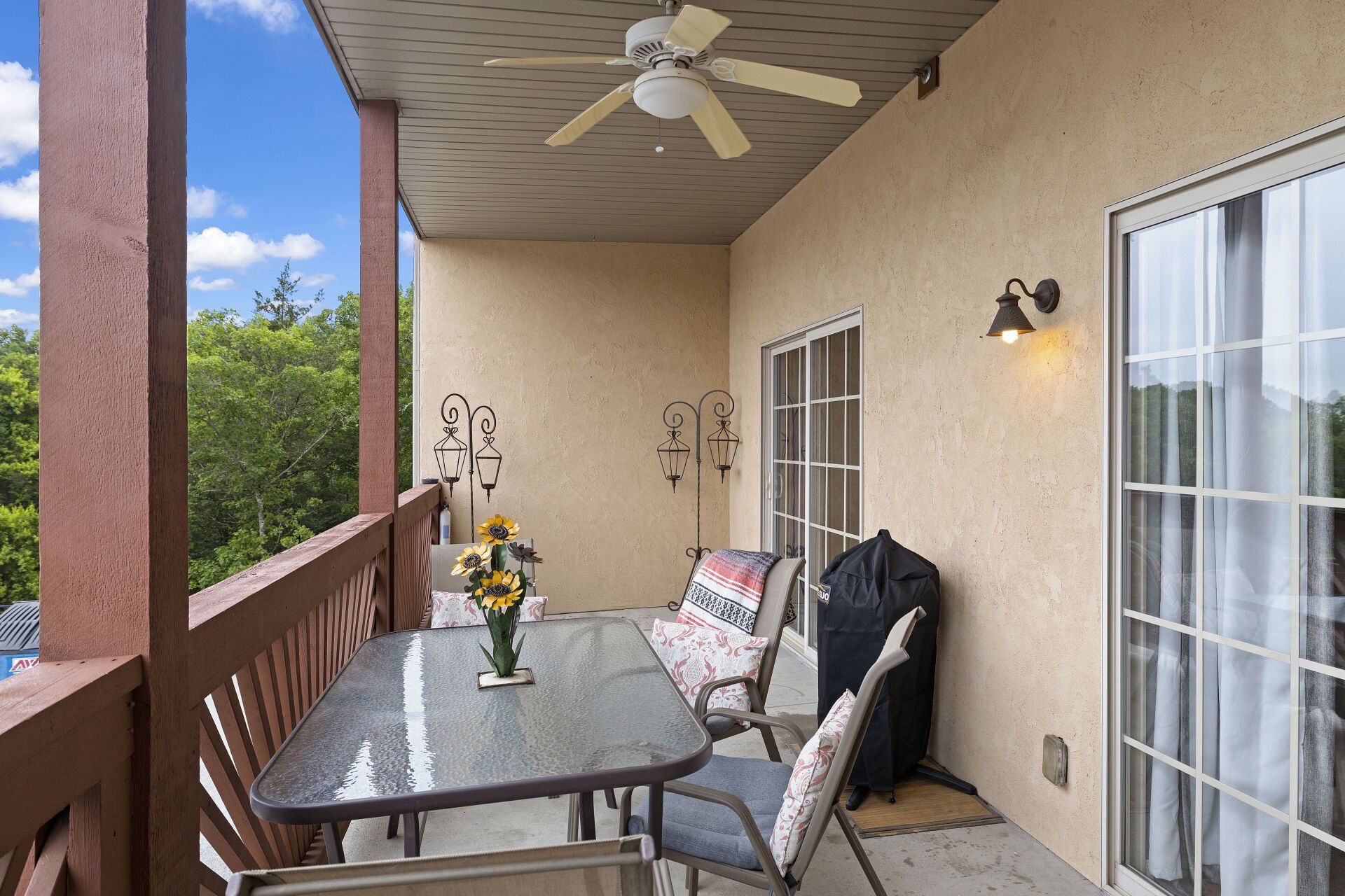 Covered patio with glass table, cushioned chairs, ceiling fan, and sliding doors beside beige stucco wall