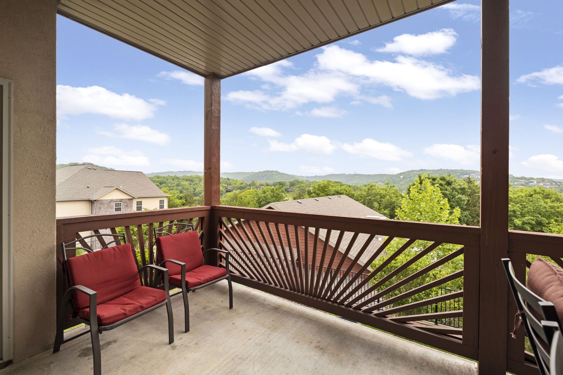 Covered balcony with red chairs, railing, and a view of rooftops and green hills under a blue sky