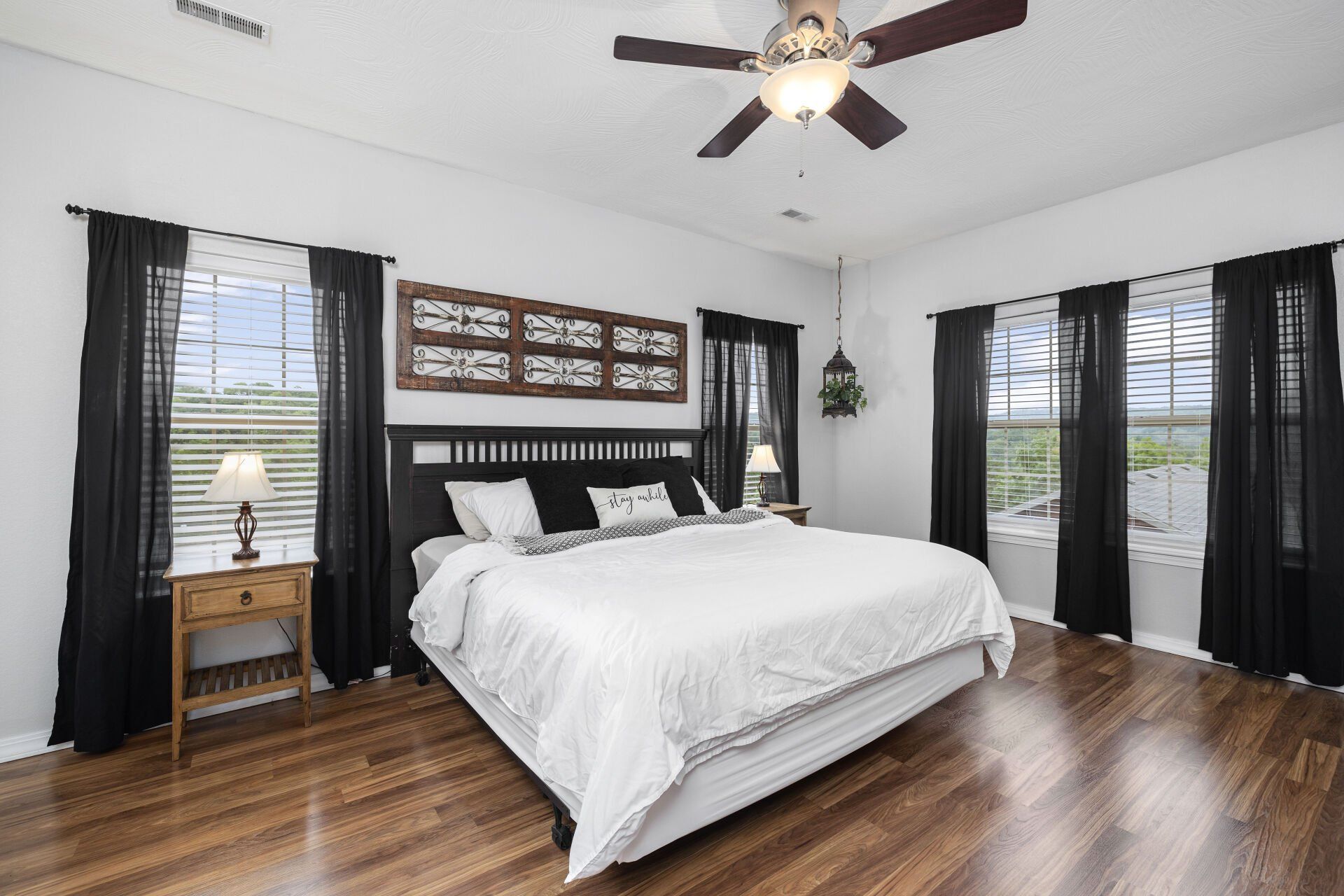 Bright bedroom with a white bed, dark curtains, wood floors, and a ceiling fan