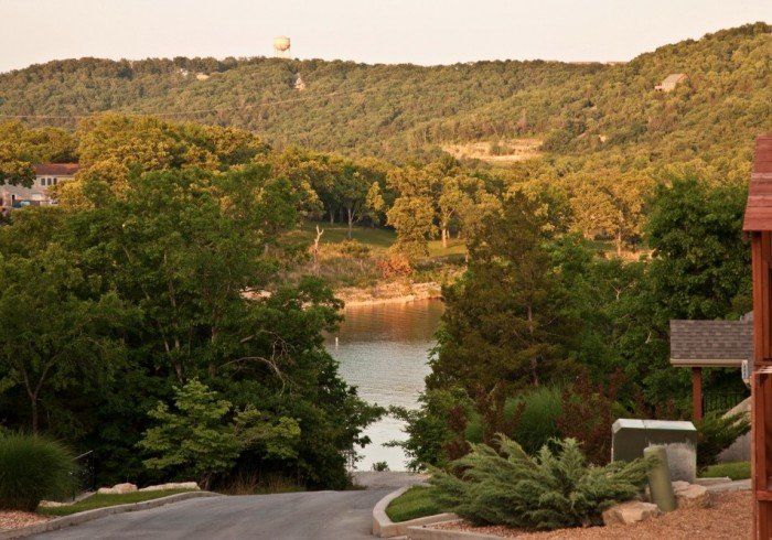 Tree-lined road leading to a calm lake, with forested hills in the background and houses at the edges.