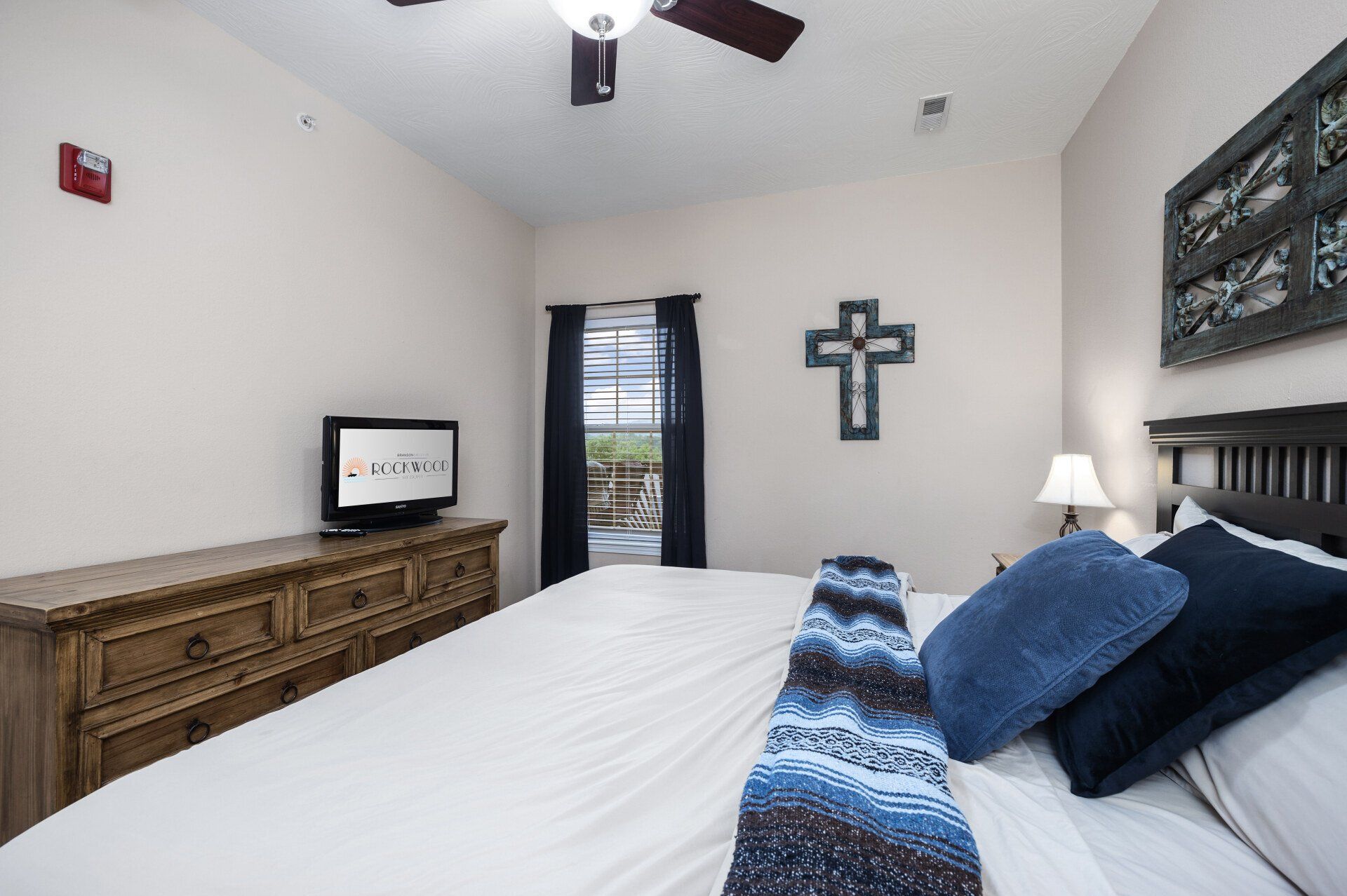 Bedroom with white bed, blue pillows, wooden dresser, TV, lamp, and cross wall decor
