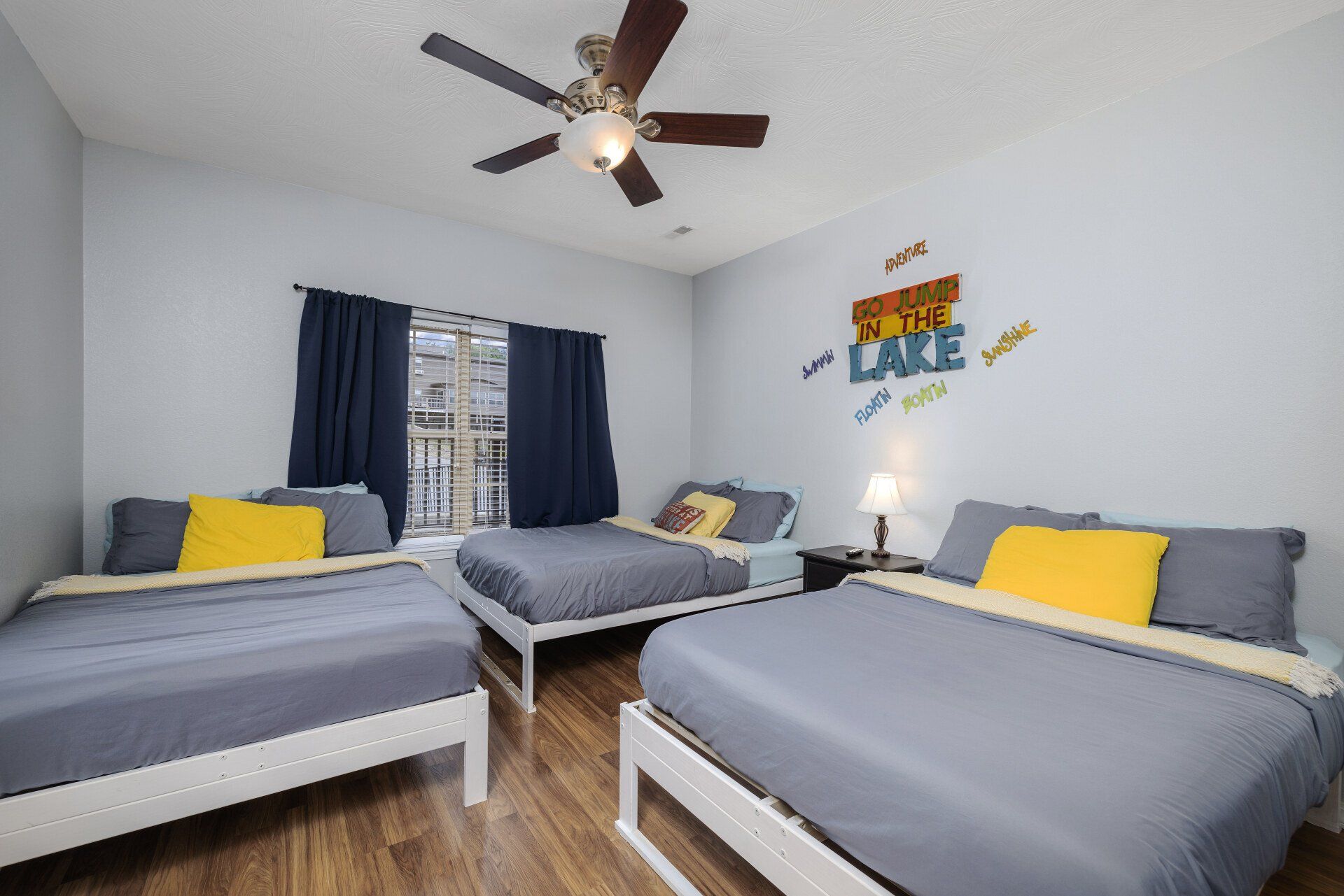 Bright bedroom with three gray beds, yellow pillows, dark curtains, and a ceiling fan