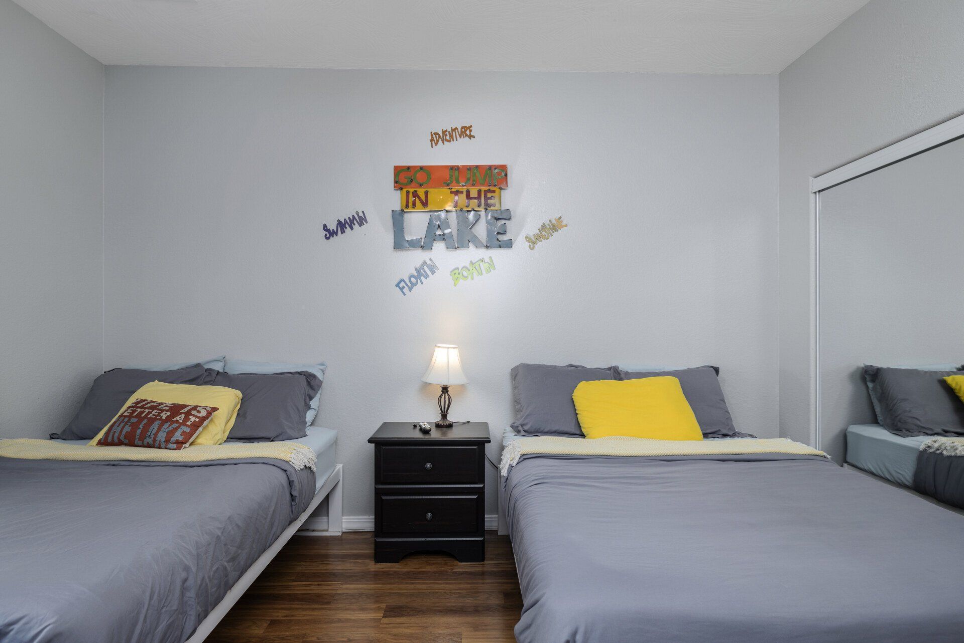 Shared bedroom with three twin beds, gray bedding, and a nightstand lamp against a white wall.