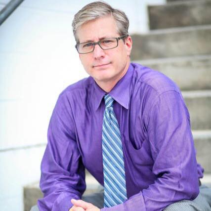 Man in a purple shirt and blue striped tie sitting on outdoor steps, looking at the camera.