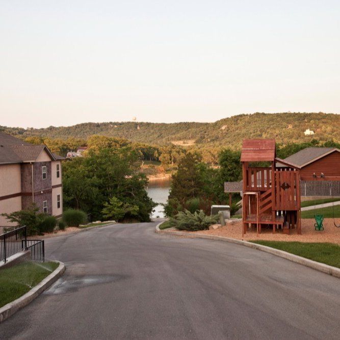 Curving road in a residential area leading toward a lake and hills at sunset, with houses and a wooden play structure