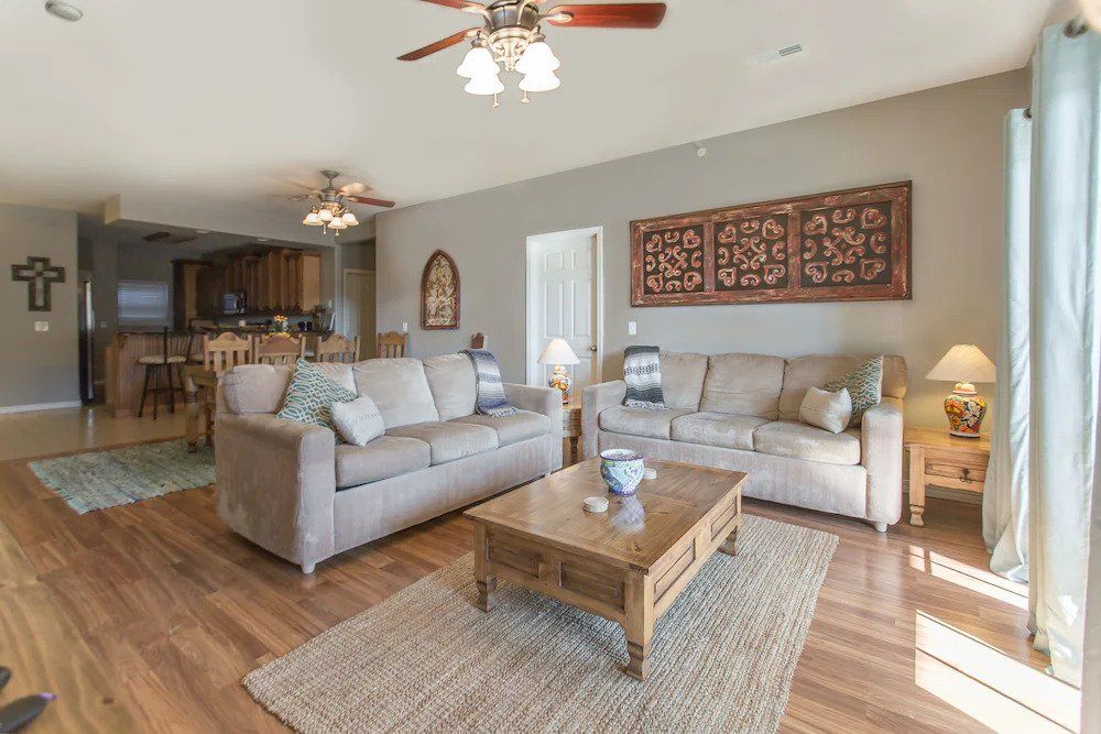 Bright living room with two gray sofas, wooden coffee table, ceiling fans, and sunlight through large windows