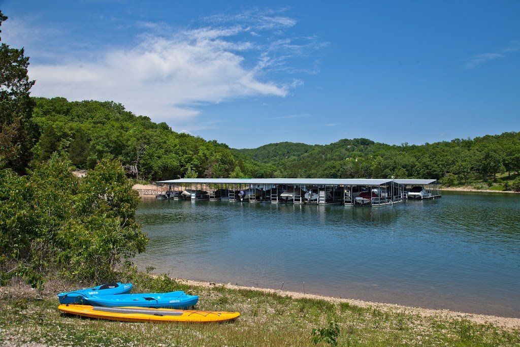 Kayaks on a lakeshore with a dock, calm water, and forested hills under a blue sky